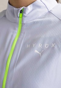 Lys grå sportsjakke med høj krave, lys grøn lynlås og reflekterende detaljer. "HYROX" logo og Puma logo præsenteret.