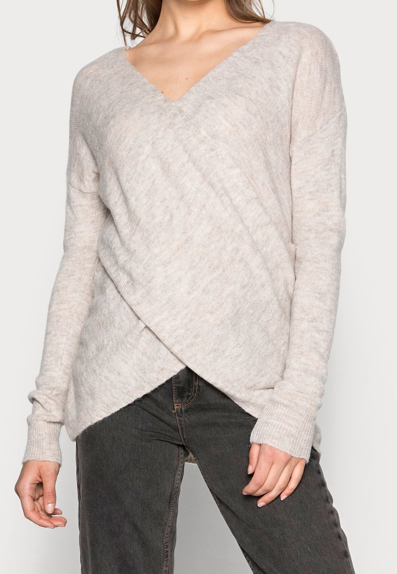 Pull en beige clair avec un col en V et un design croisé diagonal, fabriqué en tissu tricoté texturé. Présente des manches longues et une coupe décontractée.