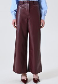 Pantaloni larghi in pelle bordeaux con una texture liscia, dotati di chiusura frontale con bottone e tasche laterali discrete, abbinati a un top azzurro chiaro.