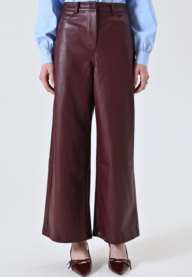 Pantaloni larghi in pelle bordeaux con una texture liscia, dotati di chiusura frontale con bottone e tasche laterali discrete, abbinati a un top azzurro chiaro.