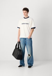 Calvin Klein hvit t-skjorte med marineblå ribbestrikket hals, blå slitte jeans og svart veske. Uformell fottøy fullfører antrekket.