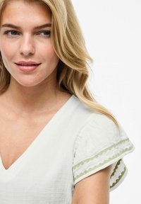 Top de algodón blanco con escote en V y mangas cortas, con bordado verde en forma de olas en los puños. Textura suave.