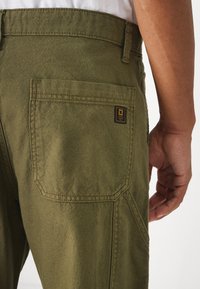 Pantalones verde oliva con una tela de algodón texturizada, que presentan un bolsillo trasero y una etiqueta de marca en amarillo y negro, vistos desde un ángulo lateral.