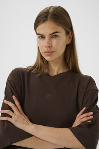 Young woman with straight brown hair wears a brown oversized shirt, arms crossed, facing forward against a plain light background.