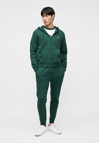 Νέος άνδρας που φοράει σκούρο πράσινο φούτερ με φερμουάρ και ταιριαστά παντελόνια φόρμας, με λευκά αθλητικά παπούτσια, σε ουδέτερο φόντο.