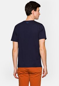 Granatowa bawełniana koszulka z okrągłym dekoltem, krótkimi rękawami i małym niebieskim logo blisko tylnego dekoltu, noszona z pomarańczowymi spodniami.