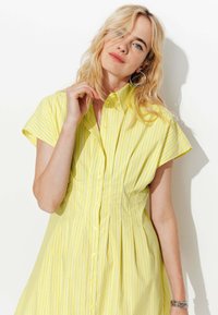 Femme blonde aux cheveux ondulés portant une robe jaune à manches courtes avec de fines rayures verticales, des boucles d'oreilles en créole et un bracelet en argent, posant devant un mur blanc.