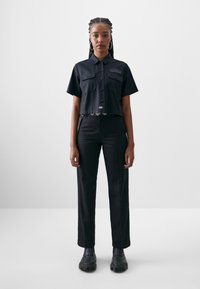 Black cropped shirt with short sleeves and two front pockets, paired with high-waisted black trousers and chunky black shoes.
