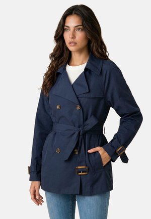 Frau trägt einen dunkelblauen, doppelreihigen Trenchcoat mit Taillengürtel und Schnallendetails, kombiniert mit hellblauen Jeans und einem weißen Top.