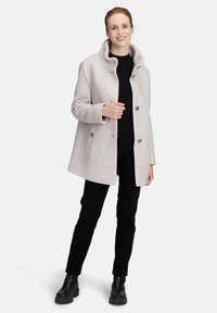 Lächelnde Frau, die einen hellgrauen, geknöpften Mantel über einem schwarzen Pullover, schwarzen Hosen und schwarzen Stiefeln trägt und vor einem schlichten Hintergrund steht.