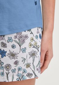 Helles blaues Oberteil mit kurzen Ärmeln, kombiniert mit einem weißen Rock im Blumenmuster, der blaue, gelbe und rosa Blumen mit grünen Stielen zeigt.