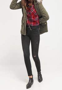 Green parka with a fur-lined hood over a red plaid shirt, paired with black slim jeans featuring a rip, and black ankle boots.