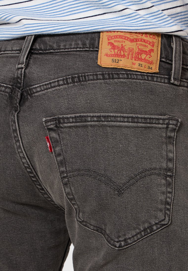 Dunkelgraue Jeans mit einer hinteren Tasche, die ein markantes, gebogenes Nähmuster aufweist. Logo-Patch am Bund angebracht; Stoffstruktur sichtbar.