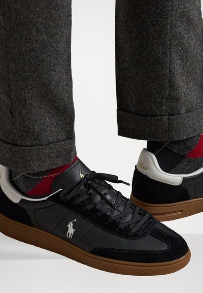 Melni ikdienas apavi, kas izgatavoti no zamšādas un ādas, ar gumijas zoli, auklu aizdari un logotipa detaļu. Apvienoti ar pelēkiem auduma biksēm un rakstainām zeķēm.