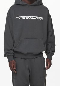 Grå huvtröja med en framficka, ribbade muddar och nederkant. Har en vit broderad "PEGADOR"-logotyp över bröstet.