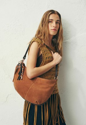 Noor naine pikkade juustega, kes kannab pruuni heegeldatud tippi ja hoiab suurt pruuni seemisnahast õlakotti tekstuurse heledava seina taustal.