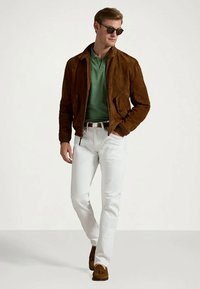 Mężczyzna w brązowej zamszowej kurtce, zielonym polo, białych spodniach, brązowych loafersach i okularach przeciwsłonecznych, idący z jedną ręką w kieszeni.