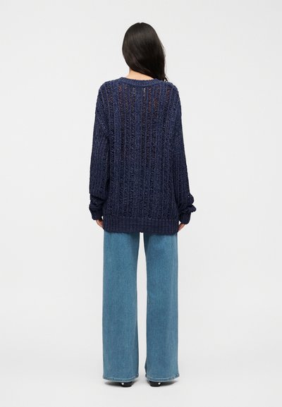 Pull en maille bleu marine avec un dos ouvert, coupe ample, encolure ronde et poignets côtelés, associé à un jean bleu à jambes larges.