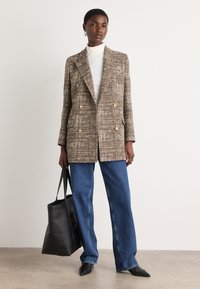 Braun karierter Blazer mit goldenen Knöpfen, kombiniert mit einem weißen Rollkragenpullover und blauen weiten Jeans. Schwarze Tote-Tasche und spitze Ballerinas.