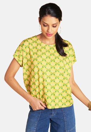 Woman with long dark hair in a ponytail wears a yellow floral short-sleeve top and blue jeans, accessorized with gold hoop earrings and bracelet.