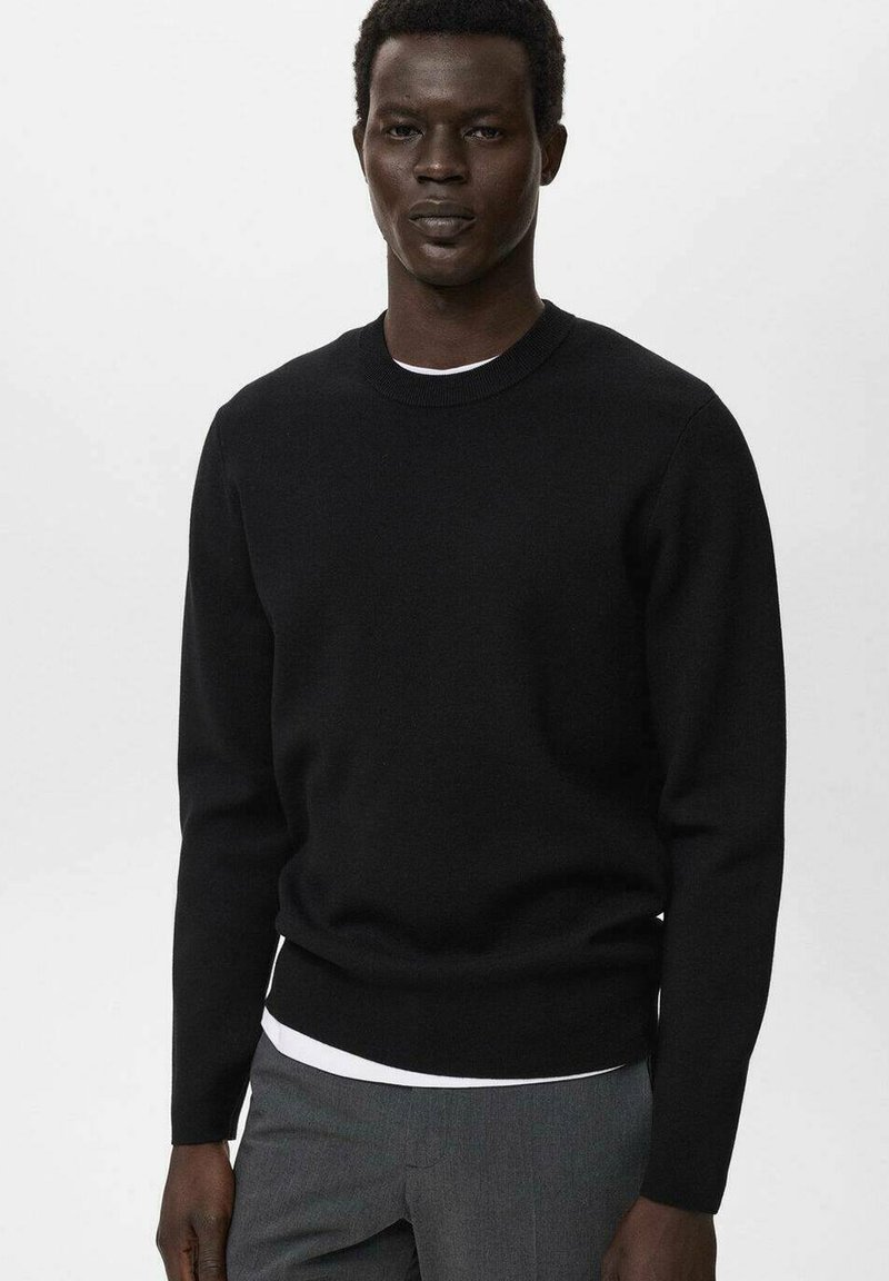 Schwarzer Rundhals-Pullover aus weichem Strickstoff, mit langen Ärmeln, gerippten Bündchen und einem taillierten Saum, über einem weißen T-Shirt getragen.