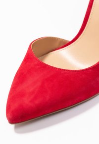 Chaussure à talon aiguille pointue en daim rouge avec semelle intérieure beige sur fond blanc, montrant l'avant et une partie du côté.