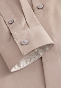 Beige skjorta med knappar och nedfällt krage, med matchande knappar och ett mönstrat foder på inre manschetten. Slät tygtextur.