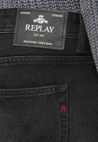 Μαύρο τζιν με δερμάτινο έμπλαστρο που φέρει την ετικέτα "REPLAY". Περιλαμβάνει μια μικρή κόκκινη κεντητή "R" στην τσέπη. Η υφή είναι λεία και ανθεκτική.