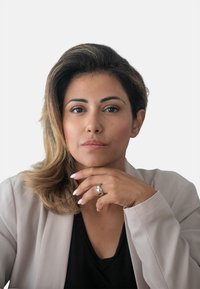 Ritratto di una persona con lunghi capelli ondulati scuri, che indossa un blazer leggero e aderente sopra una maglietta nera. Tocca il proprio mento con una mano che indossa un anello d'argento.