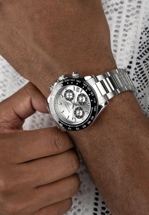Silver chronograph watch with a black bezel, three sub-dials, and a stainless steel band, worn on a wrist against a textured background.