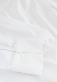Chemise blanche à manches longues avec poignets boutonnés, en tissu lisse. Caractérisée par un col classique, des manches soigneusement repliées et un design sobre.