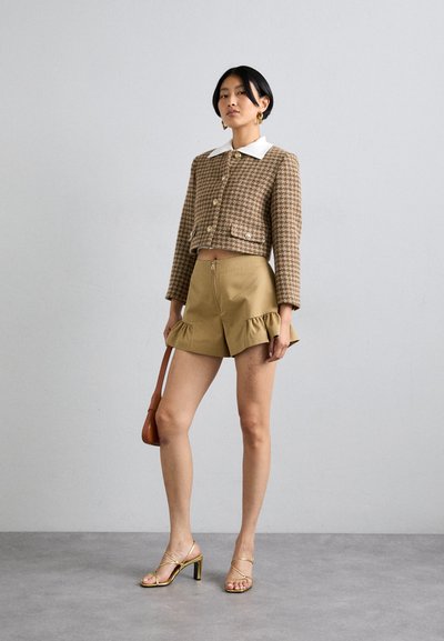 Chaqueta corta a cuadros marrones con cuello blanco, combinada con pantalones cortos de volantes khaki y sandalias doradas de tacón. La modelo sostiene un bolso marrón.