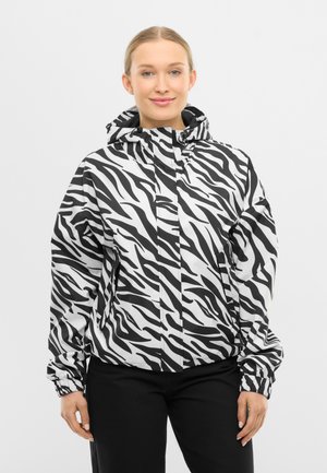 Jonge vrouw die een zwart-wit zebra gestreepte kapjasje draagt met een zwarte broek, staand en glimlachend tegen een effen achtergrond.