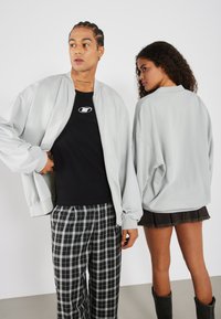 Svijetlosiva prevelika zip-jersey jakna s rebrastim manžetama, nošena preko crne majice. Modelove karirane hlače imaju crno-bijeli uzorak.