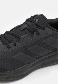 Acercamiento de una zapatilla deportiva negra con tejido de malla, superposiciones sintéticas texturizadas, cordones negros y una suela de goma con diseño.