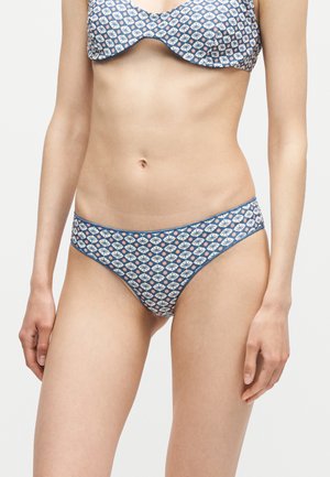 Femme portant un bas de bikini et un haut assorti bleu et blanc à motif floral, debout devant un fond clair uni.