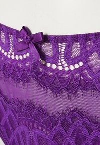 Culotte en dentelle violette avec un nœud décoratif, des motifs complexes, un tissu transparent et des bords festonnés au niveau de la taille et des jambes.