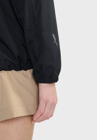 Veste noire légère avec des poignets élastiques, dotée d'un détail logo argenté sur la manche. Le matériau semble lisse et résistant à l'eau.