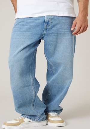 Homme portant un jean bleu clair ample et des baskets beige et blanches, debout jambes croisées et une main visible.