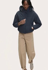 Marineblaue Steppjacke mit Kapuze, kastigem Schnitt, gesteppter Textur und Fronttaschen, kombiniert mit beigen Hosen und Sneakers.