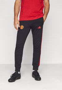 Calças de desporto pretas com riscas laterais vermelhas, apresentando o logo do Manchester United na coxa esquerda e punhos em malha.