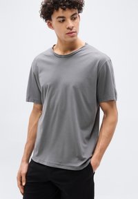 Νεαρός με κατσαρά μαλλιά, φορώντας απλό γκρι κοντομάνικο μπλουζάκι και μαύρο παντελόνι, στέκεται με το ένα χέρι στην τσέπη, κοιτάζοντας προς τα αριστερά.