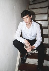 Joven con camisa blanca y pantalones negros sentado en escaleras de madera, sosteniendo una copa de vino con vino blanco, mirando a la cámara.