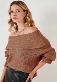 Suéter de punto marrón acanalado con escote amplio, tejido texturizado y mangas voluminosas, combinado con una falda de cuero negra.