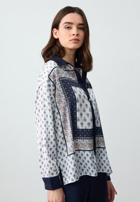 Blouse à manches longues en tissu léger et soyeux, avec une base blanche, des accents marins, des motifs complexes et un col.