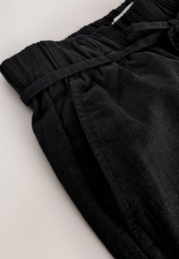 Czarne lniane spodnie z elastycznym marszczonym pasem i materiałowym paskiem zawiązanym na supeł, pokazujące szczegół bocznej kieszeni.