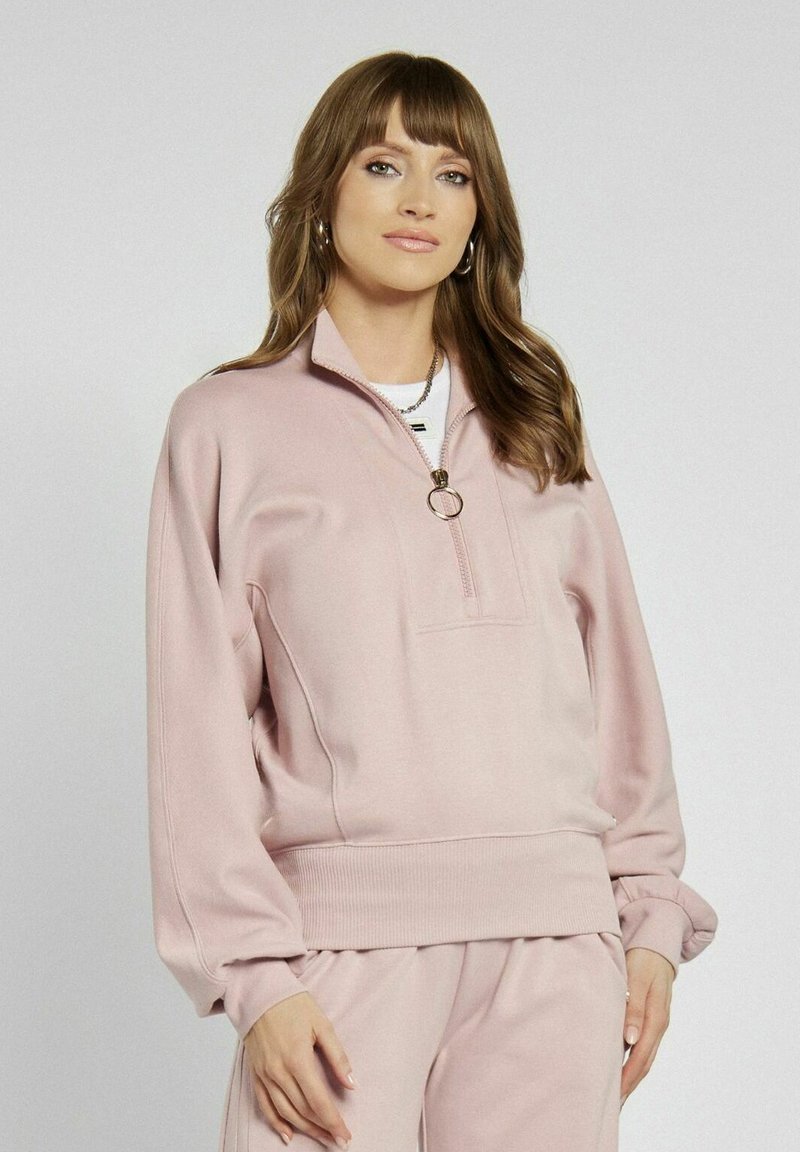 Jasnoróżowy sweatshirt z półzamkiem, ściągaczem na dole i miękkim materiałem. Posiada przednią kieszeń oraz szerokie rękawy dla luźnego kroju.