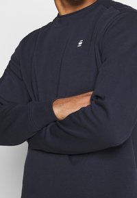 Marinblå sweatshirt i mjukt tyg med rund hals, ribbade muddar och en liten vit logotyp på bröstet.