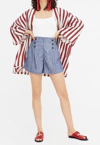 Kimono léger à rayures avec des manches larges en rouge et blanc, associé à un short taille haute à rayures bleu avec boutons sur le devant. Sandales rouges.