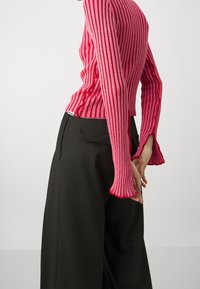 Różowy sweter z prążkowanego materiału, dopasowany w ciele, z rozszerzanymi rękawami i falbaniastym dołem. W połączeniu z czarnymi spodniami typu wide-leg.